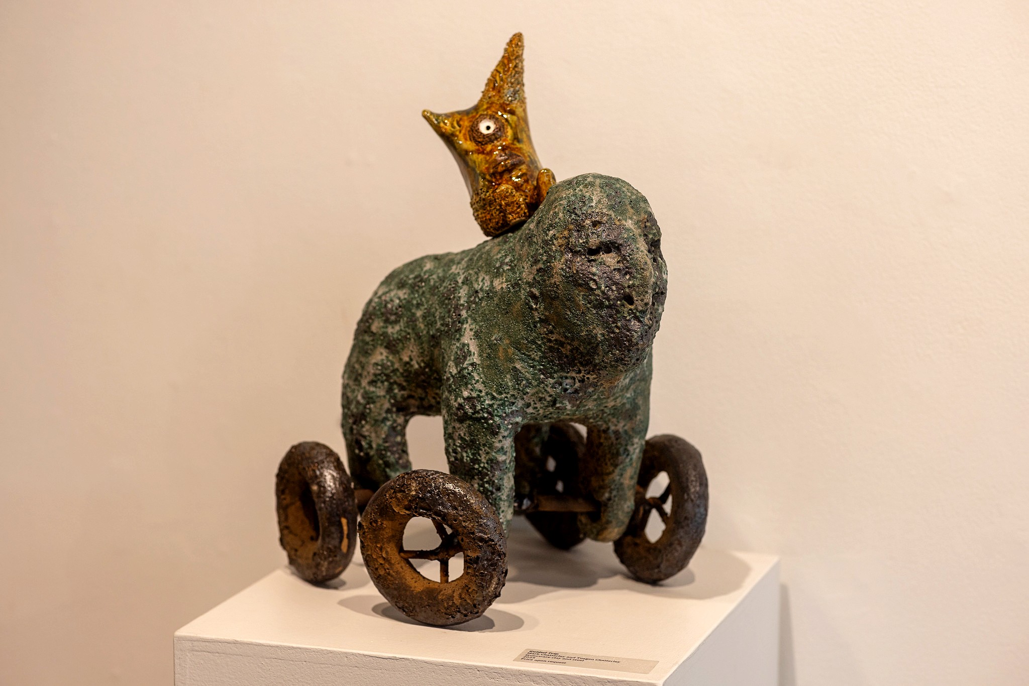 A ceramic sculpture of a green creature on four rust-colored wheels displayed on a white pedestal. Atop the creature sits a small, textured yellow and brown figure with wide eyes and pointed ears.