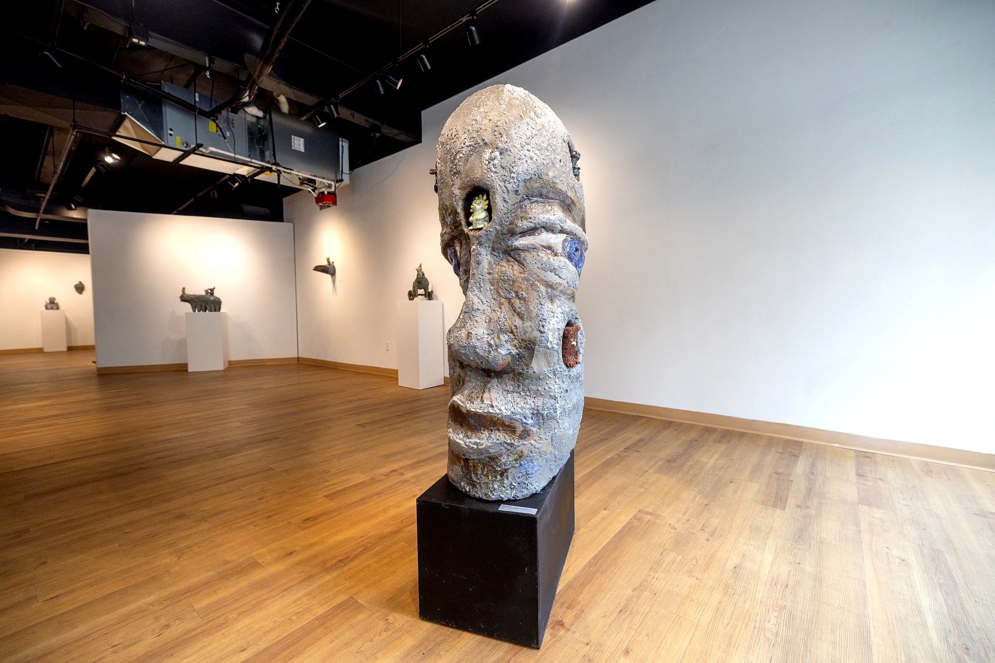 A large ceramic sculpture of a human head on a black pedestal in a gallery. The head is textured with exaggerated facial features and small, colorful creatures embedded in its surface.