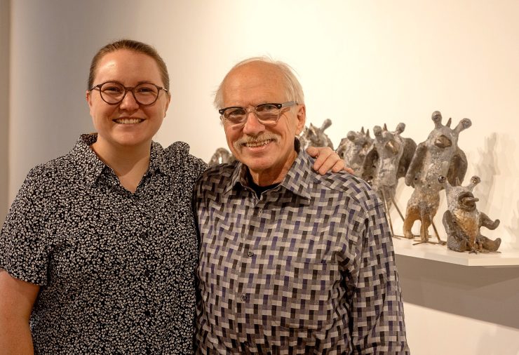 Two people stand smiling in a gallery. The person on the left has their arm around the person on the right. To the right is a shelf full of small, creature-like sculptures.