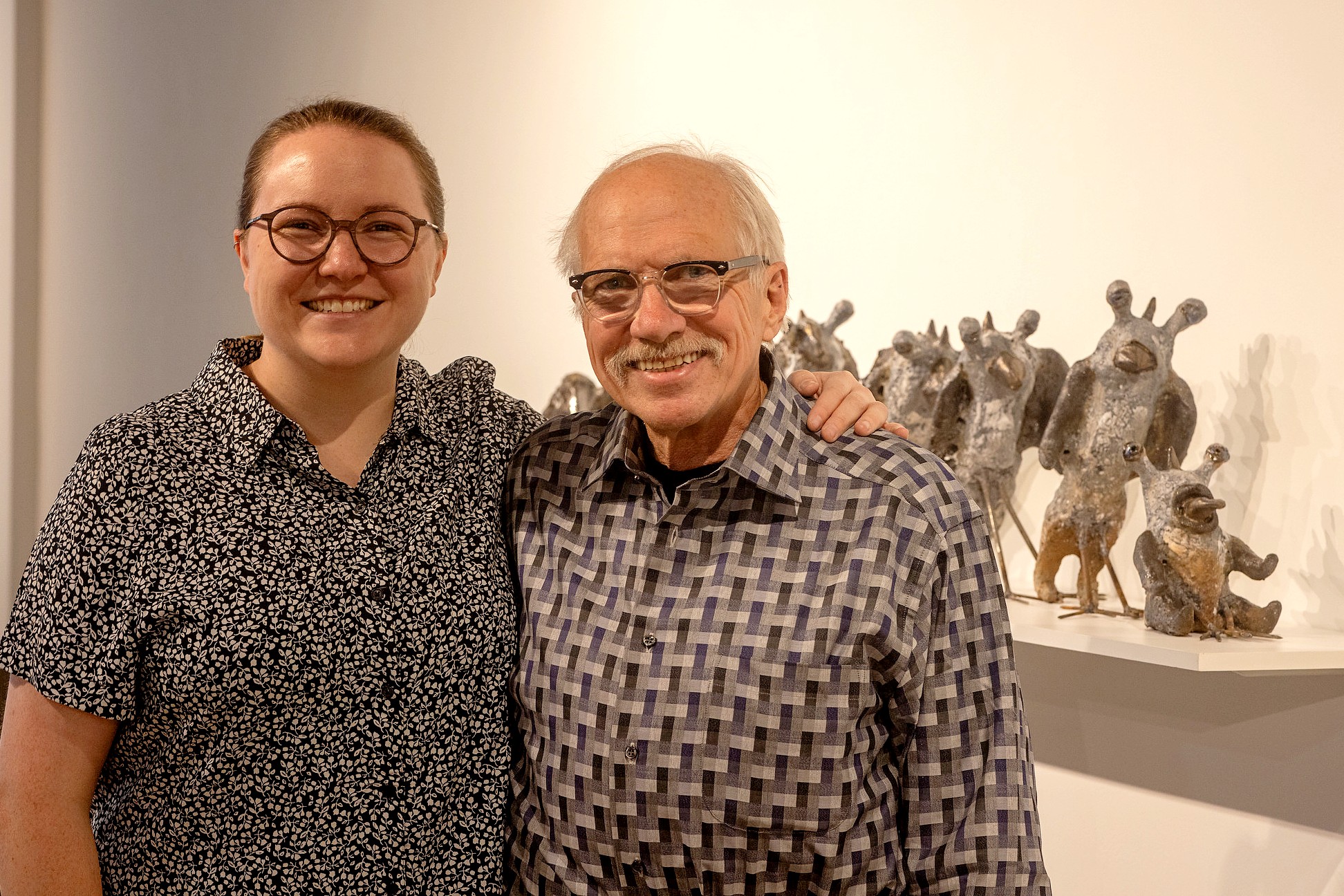 Two people stand smiling in a gallery. The person on the left has their arm around the person on the right. To the right is a shelf full of small, creature-like sculptures.