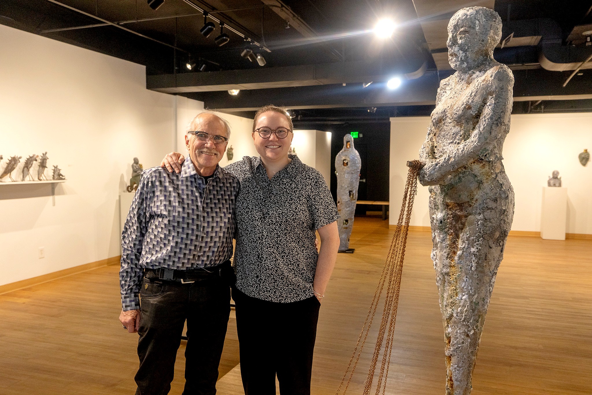 Two people standing smiling in a contemporary gallery. The person on the left has their arm around the person on the right. To the right is a tall, textured sculpture of a human figure holding chains connected to small, creature-like figures on the floor.