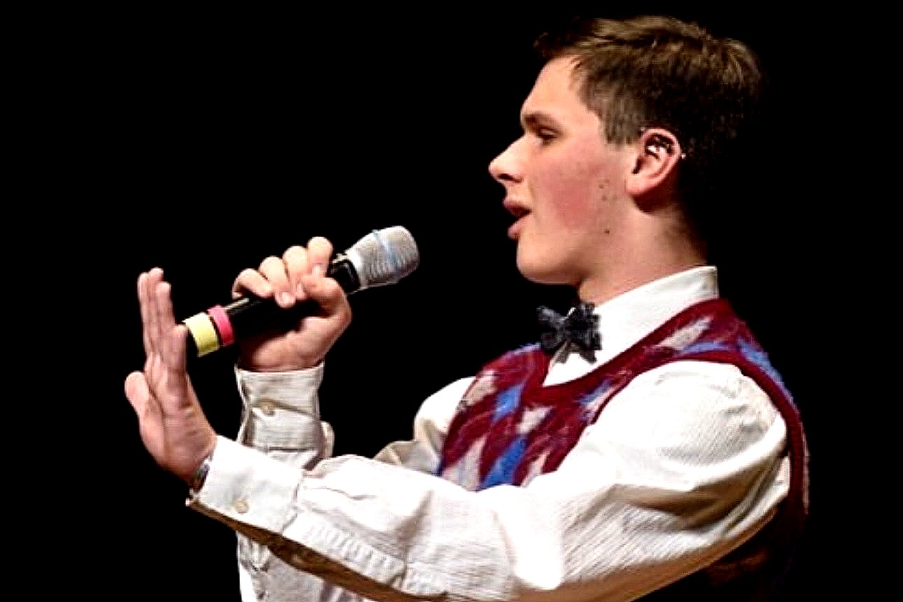 A photo of an individual performing on stage, holding a microphone and wearing a patterned sweater vest with a bowtie. The background is dark, emphasizing the performers focused expression.