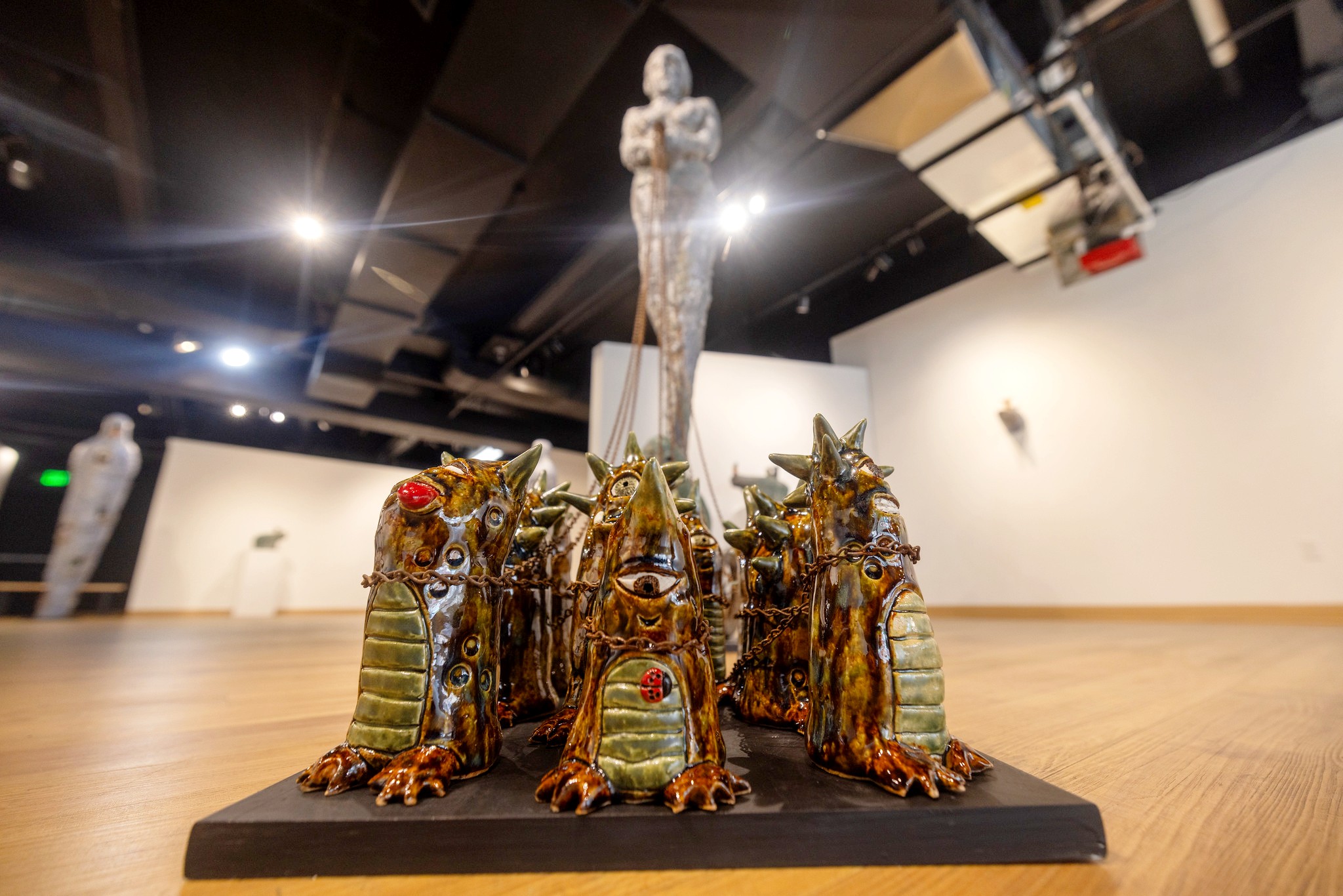 Low-angle view of a cluster of whimsical, glossy ceramic creatures arranged on a black base. Each has a horned, reptilian form with chains draped across their bodies. The background shows a tall figure sculpture holding the chains.