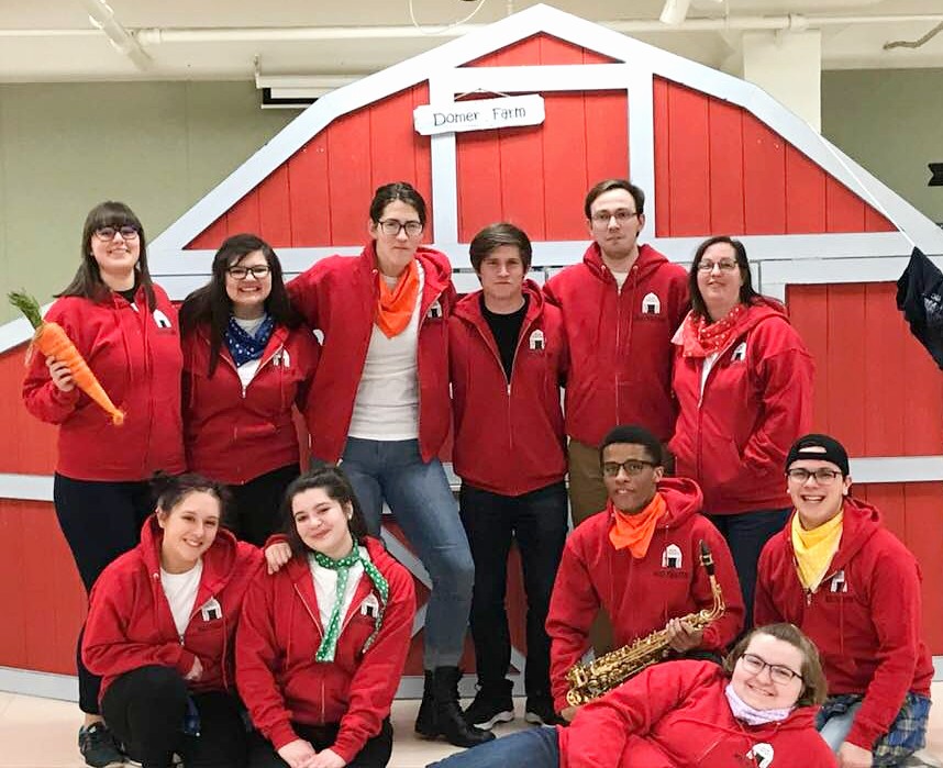 Eleven people all wearing red hooded sweatshirts standing and sitting in front of a red barn theatre set and on the barn there is a sign that says 