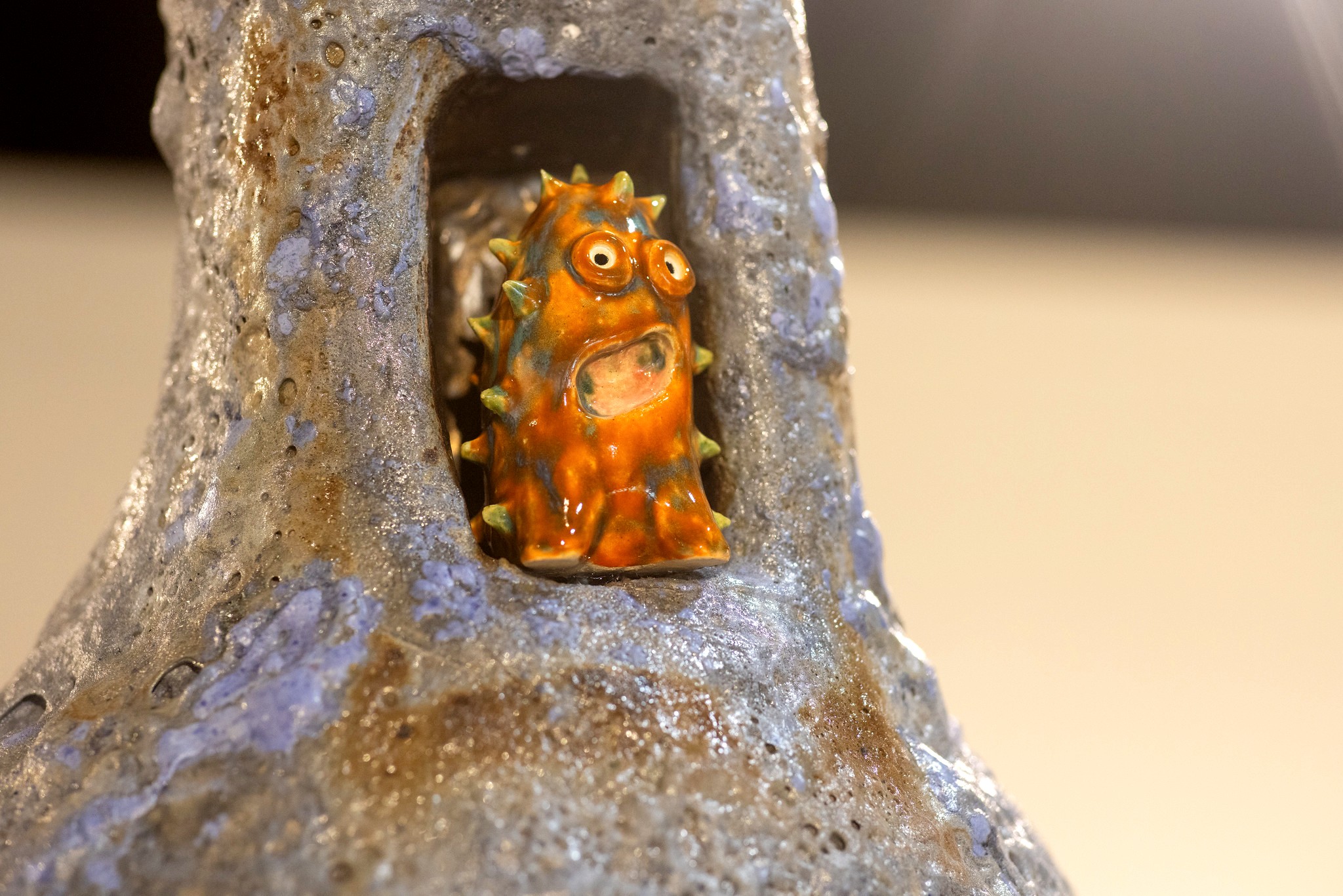 A close-up of a bright orange, spiky ceramic creature with large eyes and an open mouth. It is nestled into an alcove of a tall, gray and blue textured sculpture.