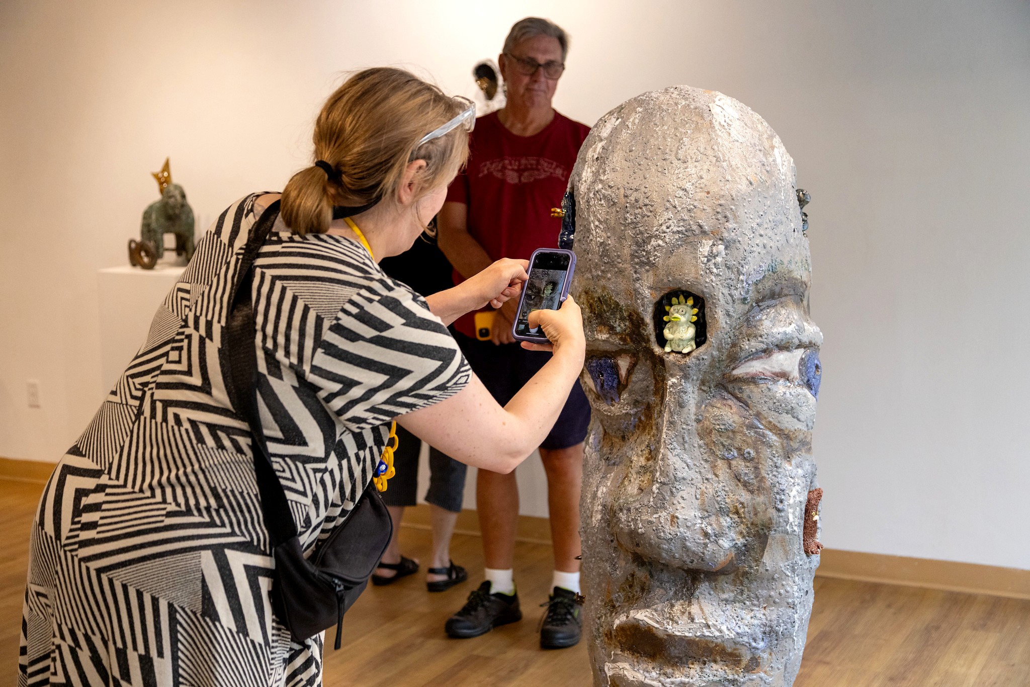 A woman taking a picture with her cell phone of a large ceramic sculpture of a human head that is textured with exaggerated facial features and small, colorful creatures embedded in its surface.