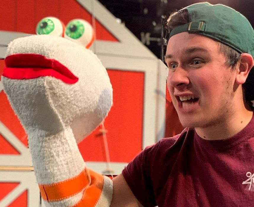 A man wearing a green baseball hat backwards and a maroon T-shirt. He is performing with a puppet on his right hand.