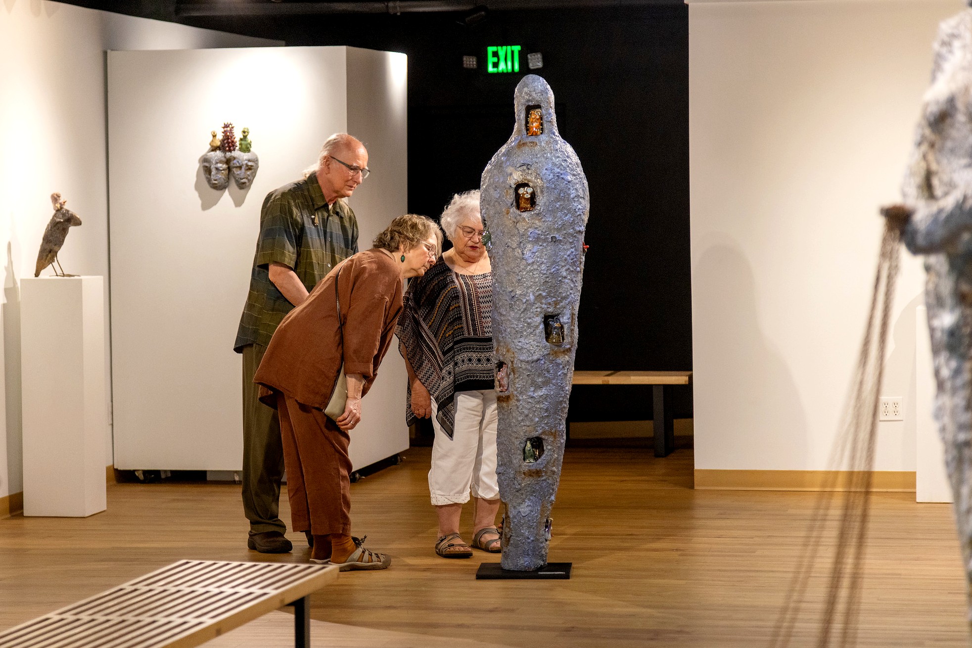 Three people, one man and two women, looking at a tall, cocoon-shaped sculpture with embedded miniature figures in caves along its surface. The piece stands upright on a black pedestal in the center of a gallery room.