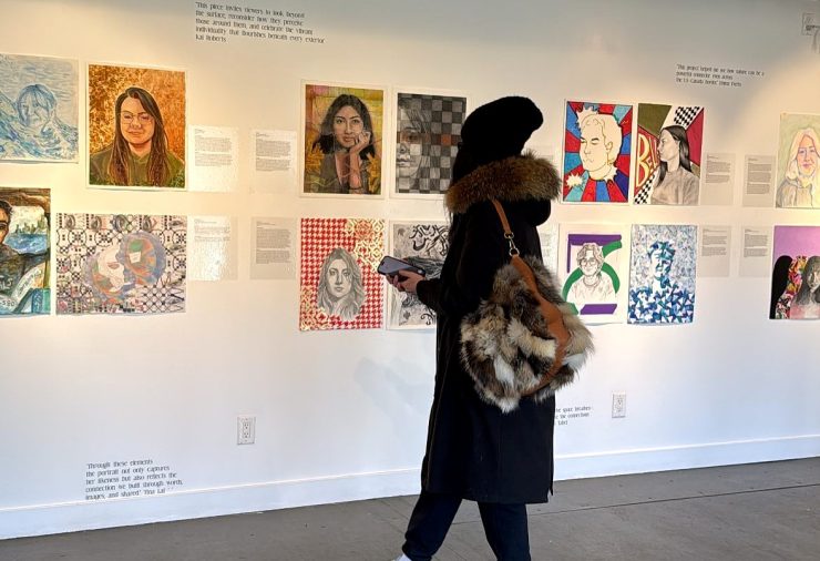 Woman in a long black winter coat and black knit hat carrying a fake fur covered purse and a cell phone looking at a wall of painted and drawn portraits at a gallery.