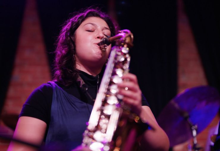 A woman wearing a black turtle neck and blue shirt plays a saxophone on a stage.