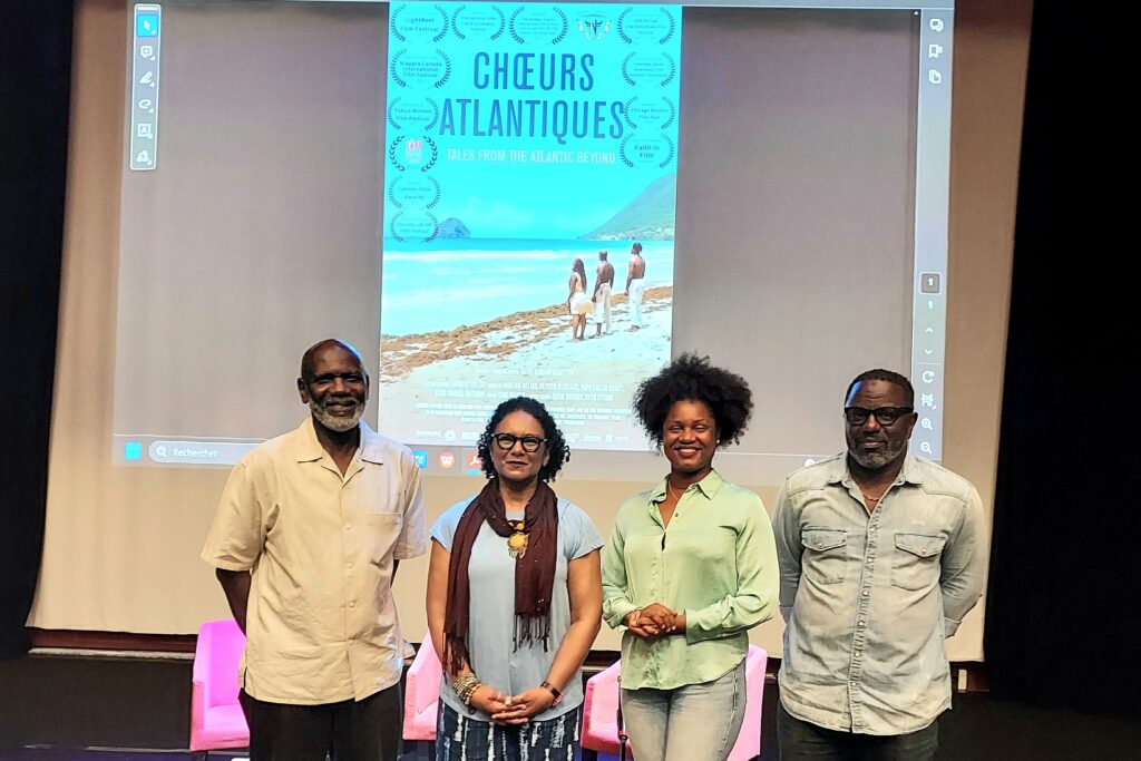 Four black people stand in a row in front of a movie screen. On the movie screen is a projected image of the