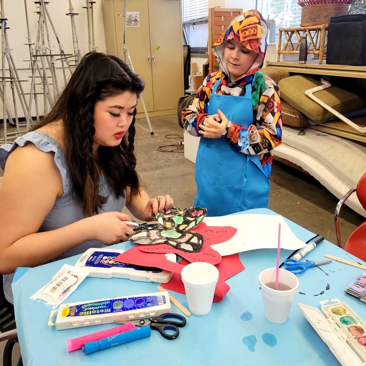 A young adult assists a child with an art project at a table covered in supplies. The child, wearing a colorful hoodie and a blue apron, watches as the adult works on a butterfly-shaped paper cutout decorated with paint and markers.