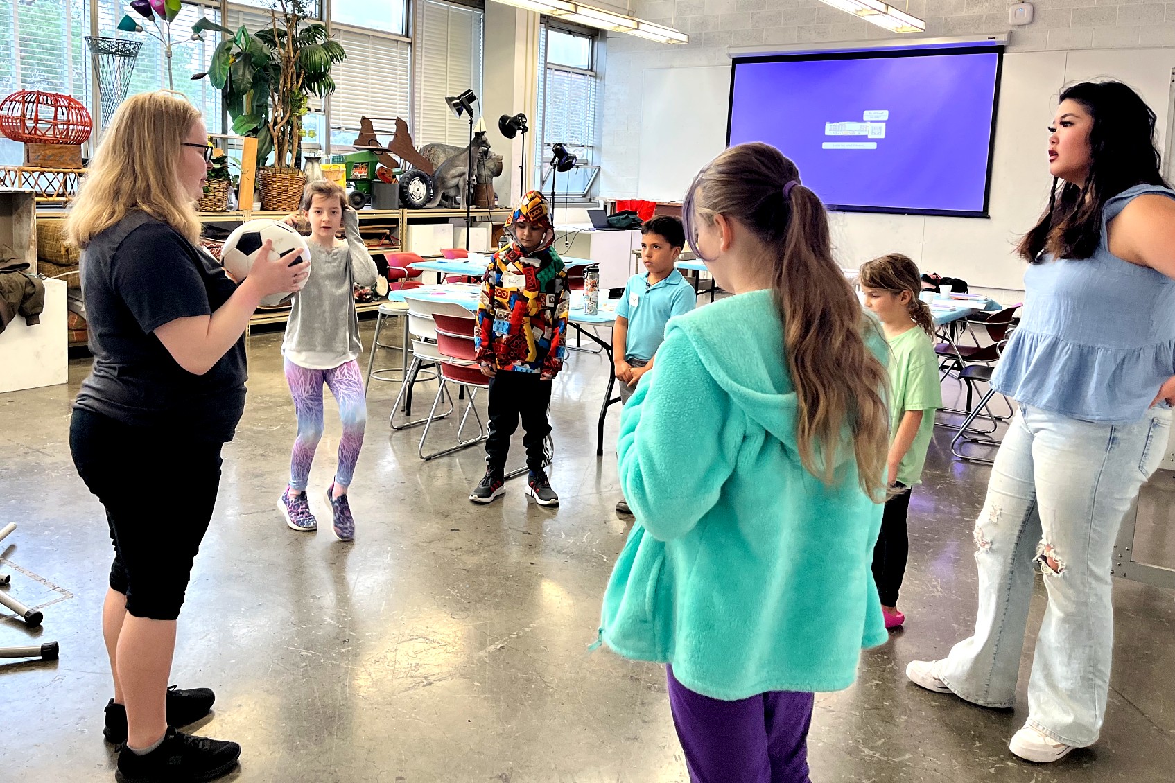 Children and two instructors stand in a circle during an activity in an art classroom. One instructor holds a soccer ball while leading the group.