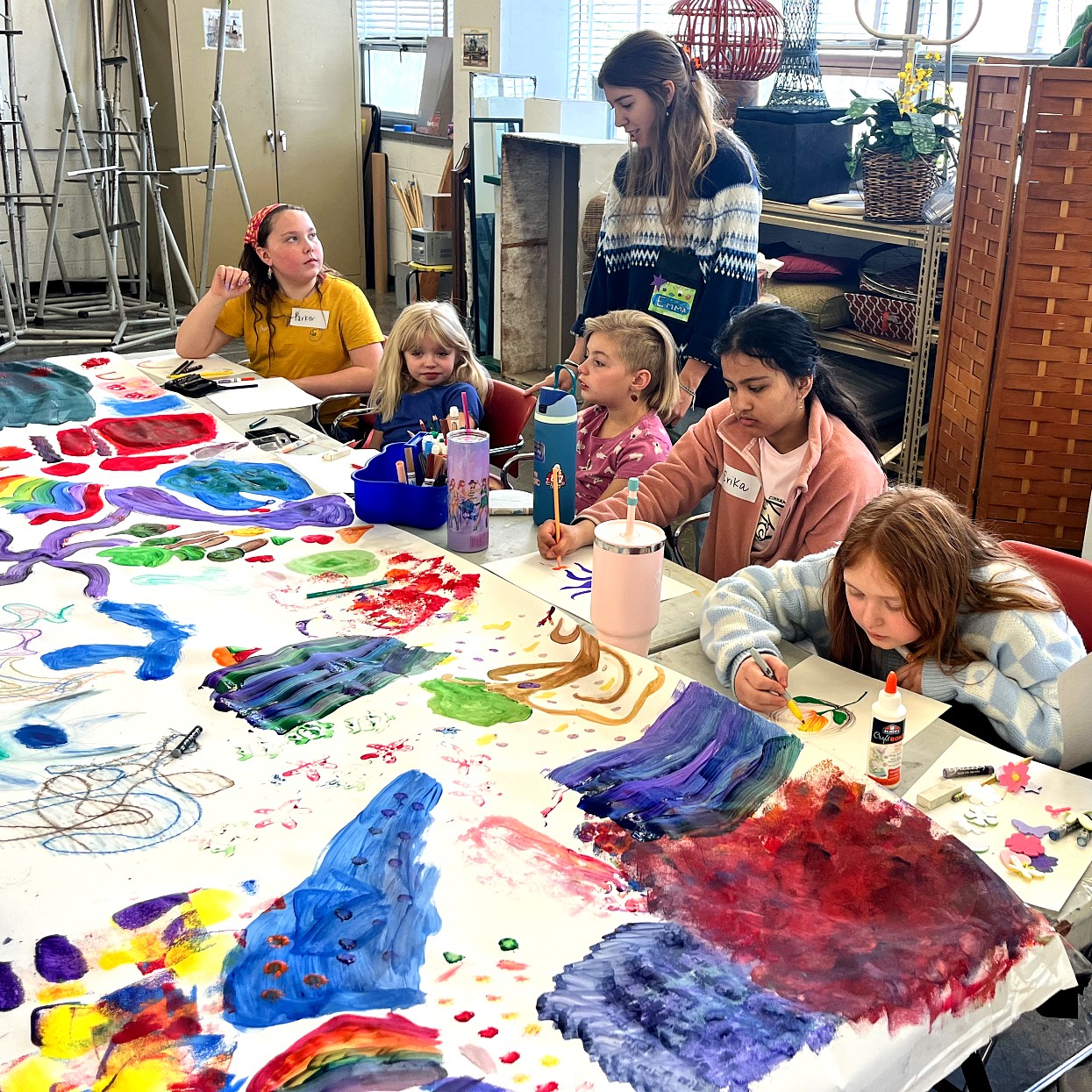 A group of children sit around a large paper-covered table, painting and drawing together. An instructor stands behind.