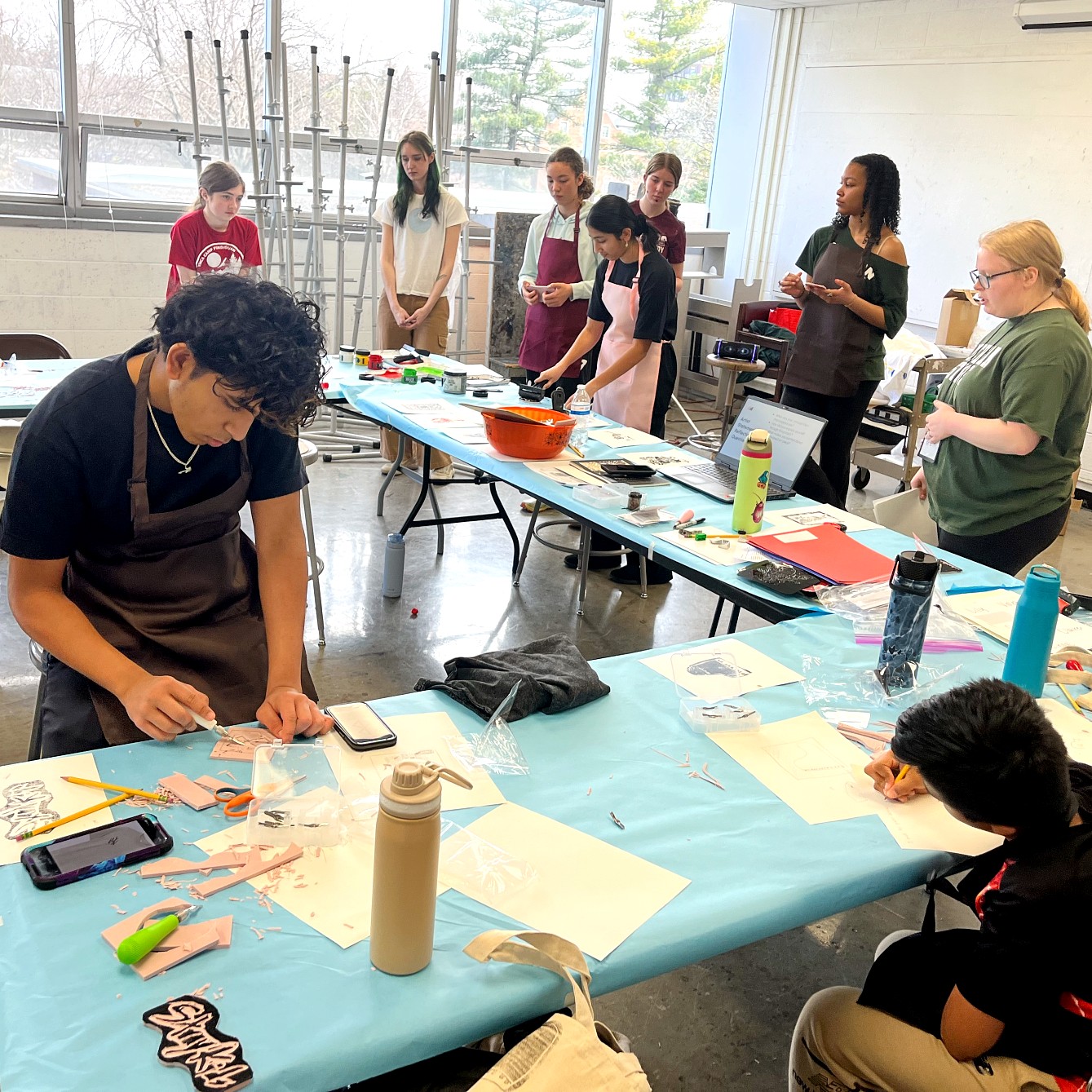 A classroom scene with students and instructors working on printmaking projects. In the foreground, a student in a brown apron carves a design into a pink block while another student sketches behind him. In the background, several students stand listening to an instructor.