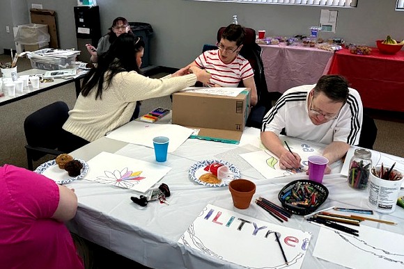 A photo of two adults at the same art workshop. One is drawing and coloring a large flower, while the other works on a design using a cardboard box as a surface. A facilitator sits nearby, offering guidance.