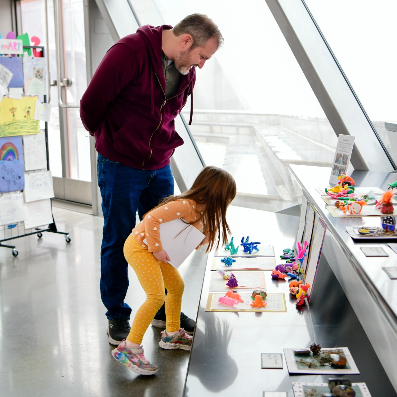 A young child and an adult bend down to closely view a display of colorful clay sculptures and artwork.