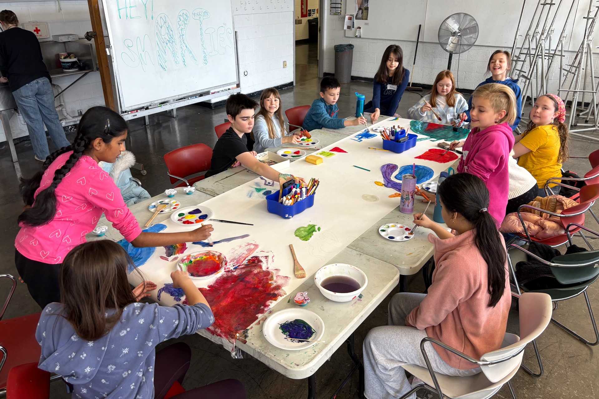 A group of about a dozen children sit and stand around a long table, painting together on a larger sheet of white paper that stretches across the shared workspace.