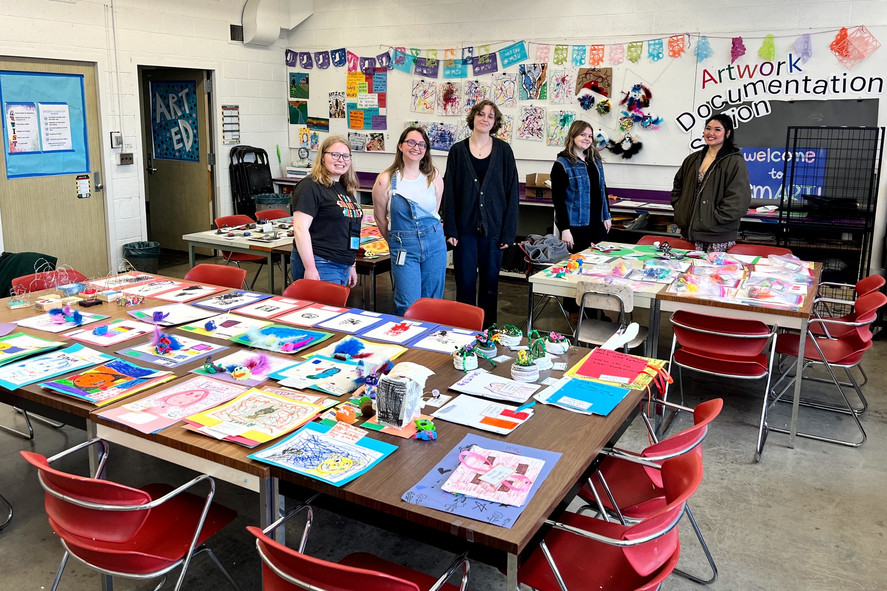 Five young adults stand smiling behind the tables covered with children's artwork in a classroom. The artwork includes bright paintings, collages, and 3D craft projects made with feathers, clay, and paper.