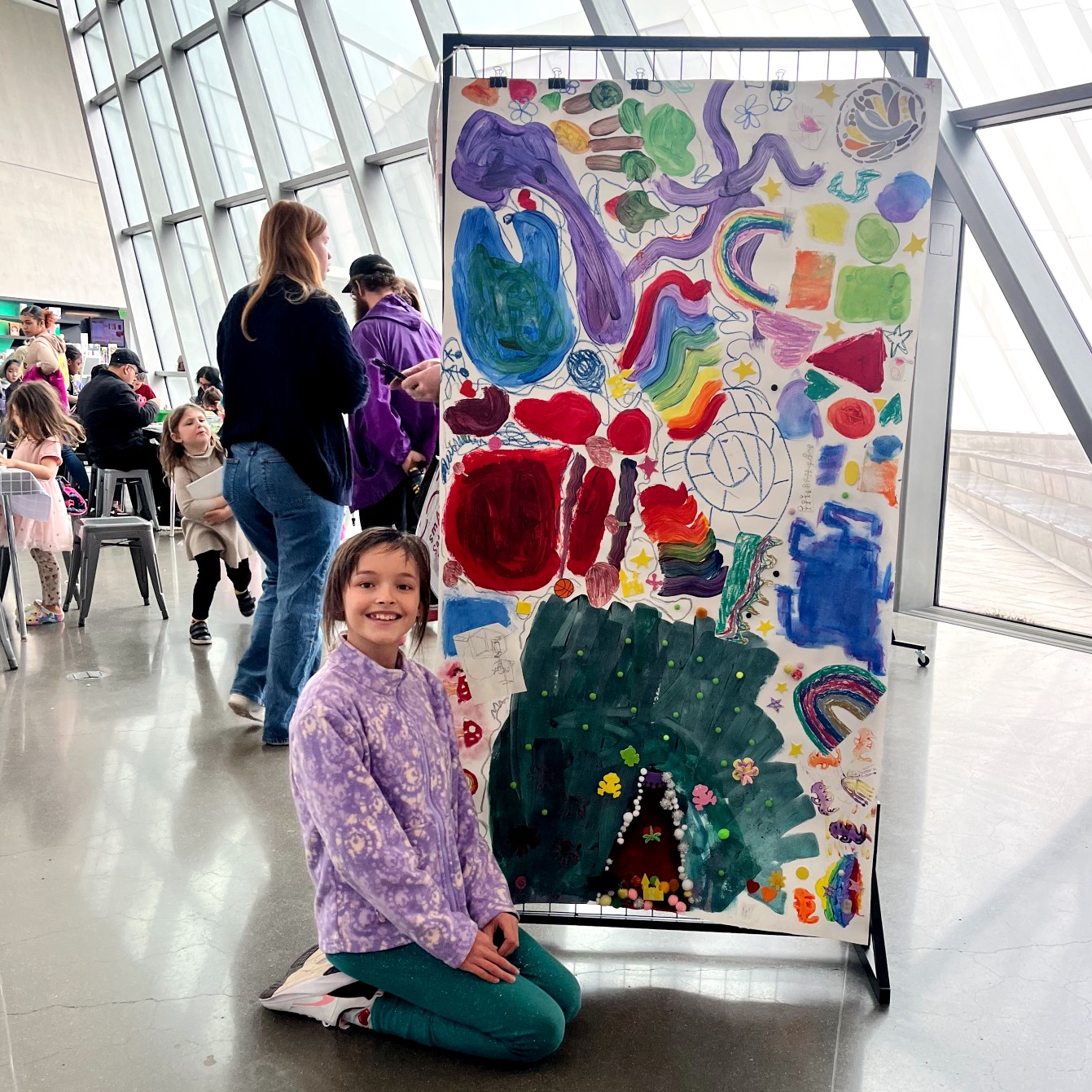 A young child kneels on the floor of a bright gallery space, smiling in front of a tall display panel covered in collaborative children's artwork.