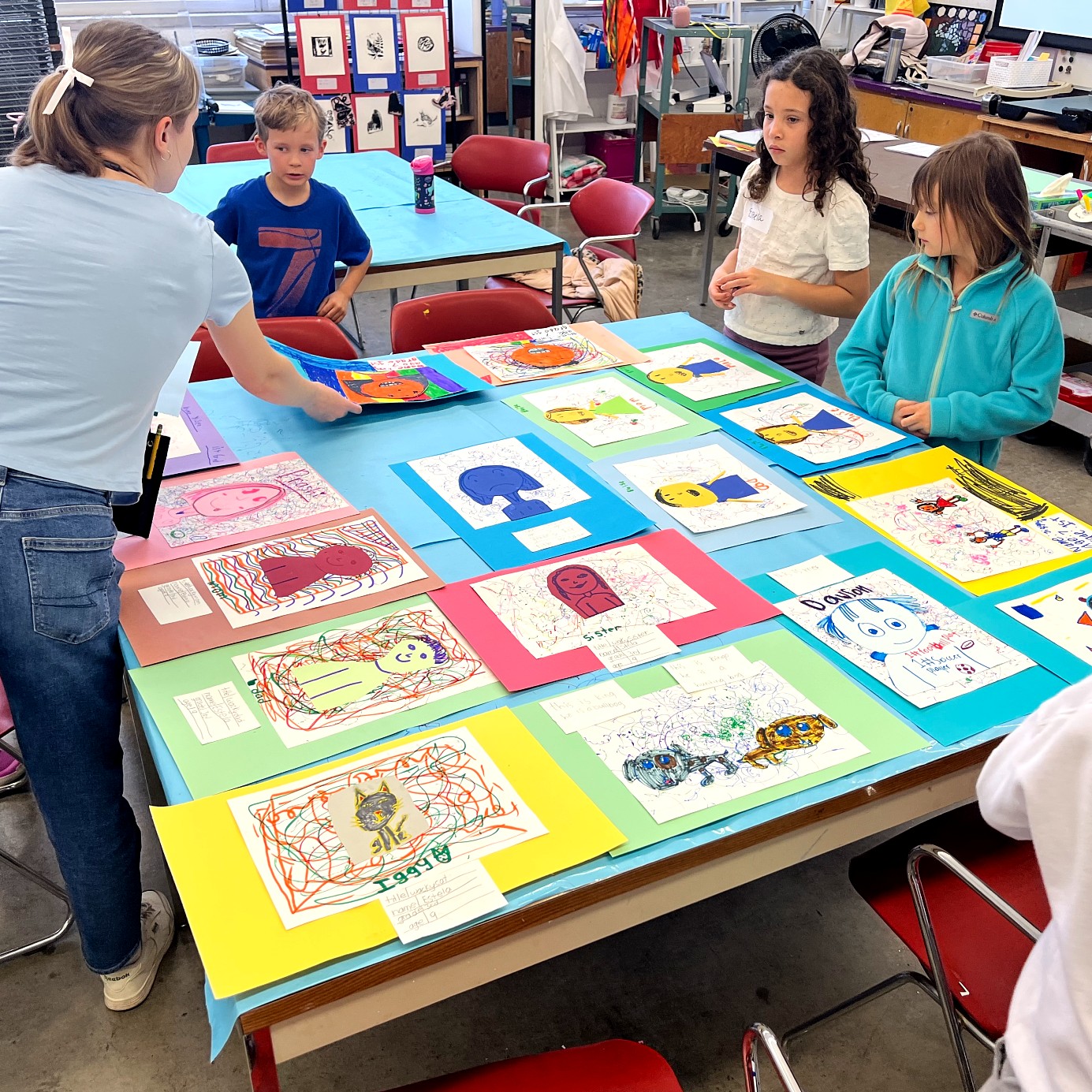 Children and an instructor gather around a table covered with completed self-portrait drawings mounted on brightly colored construction paper.