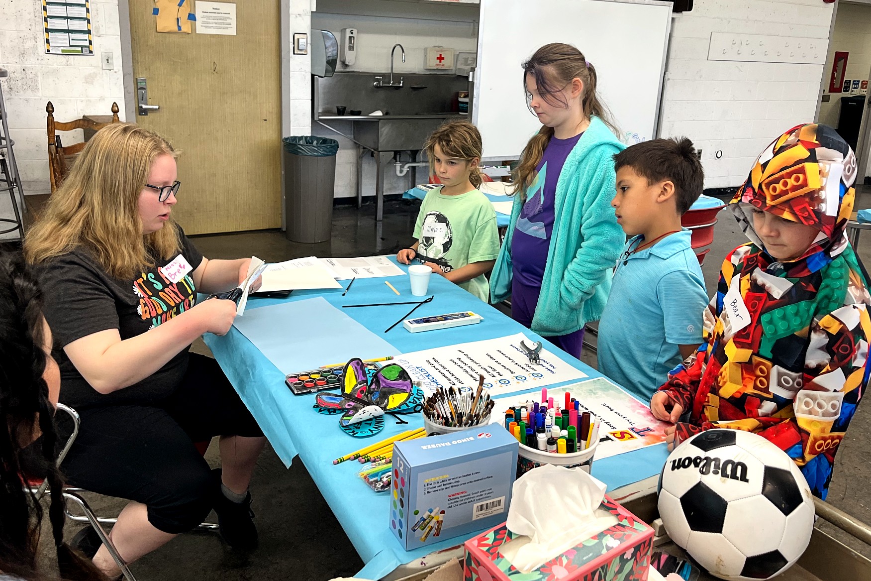 An instructor with scissors demonstrates a cutting technique at a table as several children look on closely.