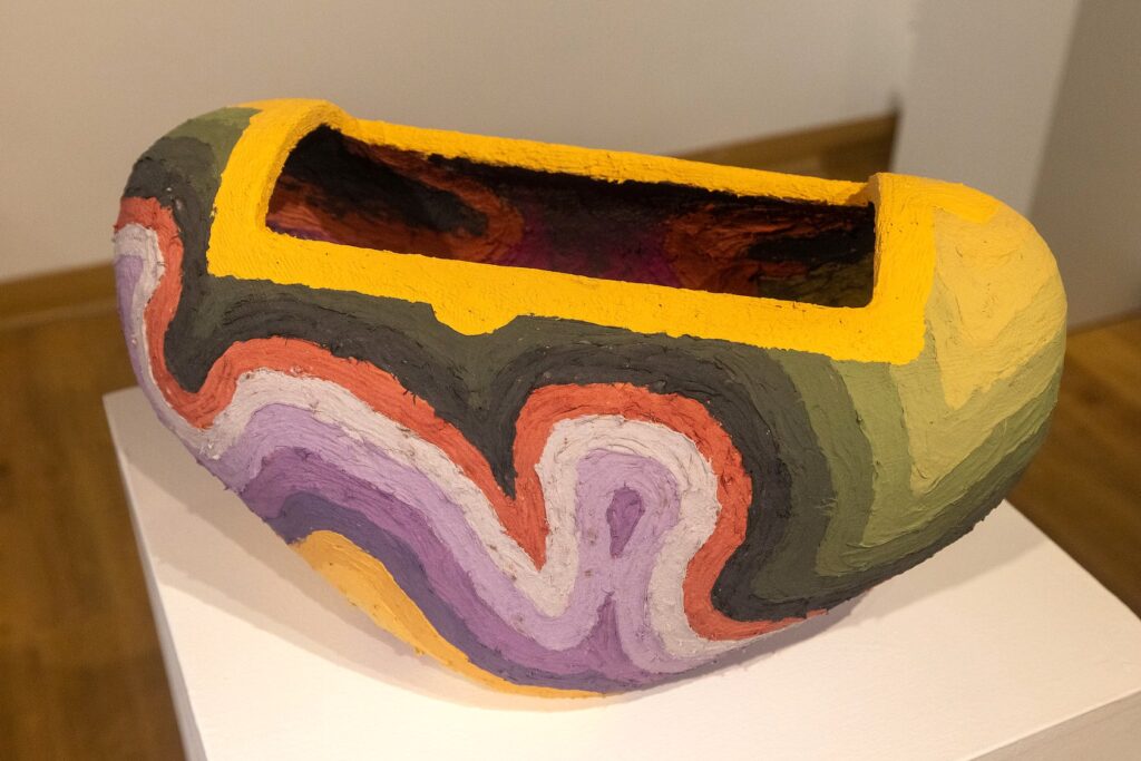 A brightly colored ceramic vessel with a wide rectangular opening sits on a pedestal. The surface features layered, wavy stripes of green, orange, red, purple, and yellow.