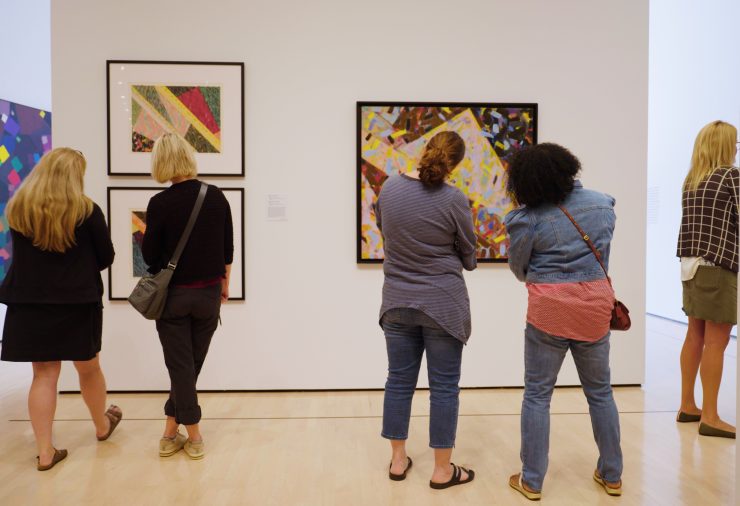 Four women stand in front of a wall of art. Two women on the right look at two paintings stacked one above the other on the wall. The other two women look at another painting on the right, their heads are tilted in towards each other.