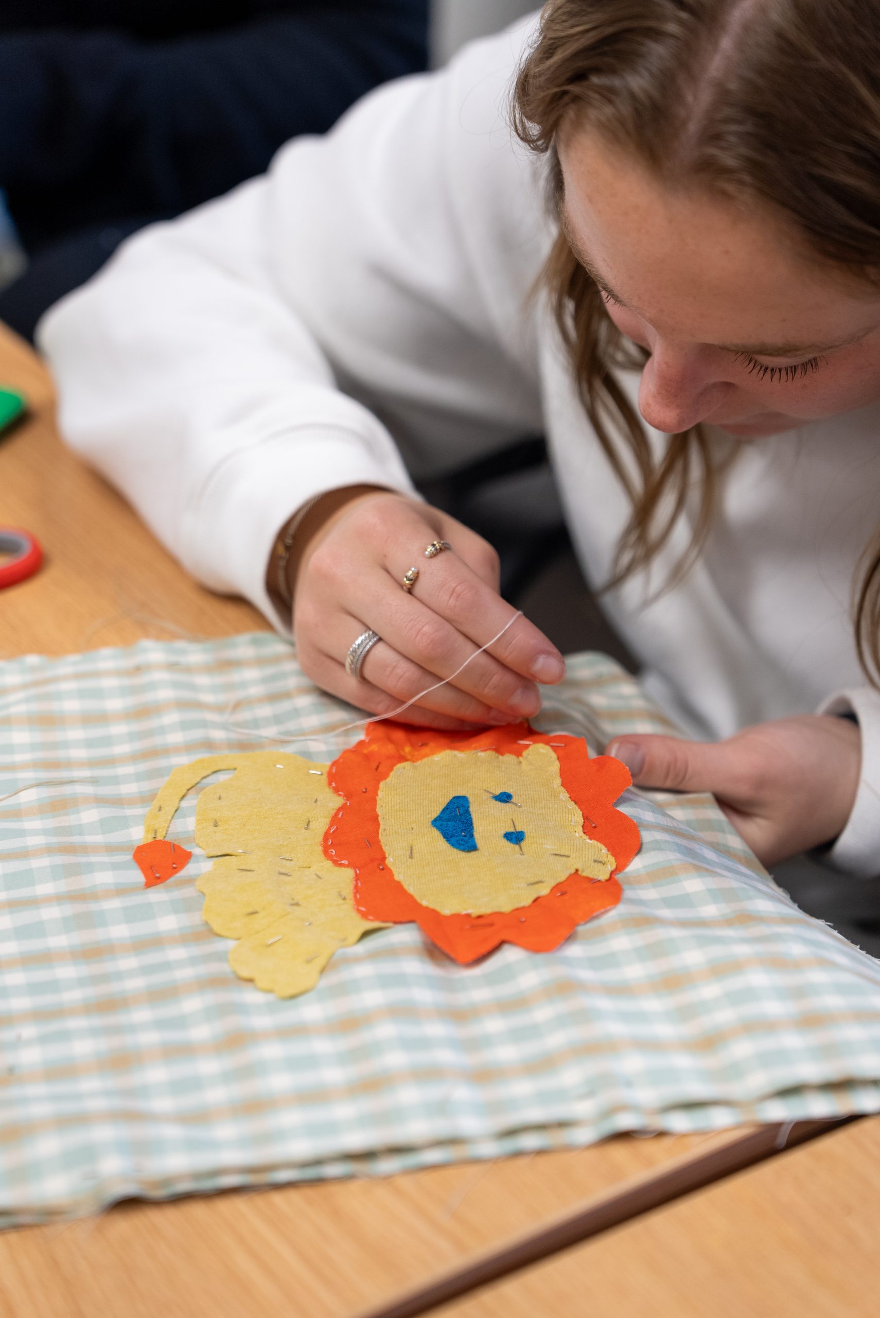 A girl wearing silver rings is leaned over a table, she holds a needle and thread sewing a cutout of a lion onto a piece of cloth.