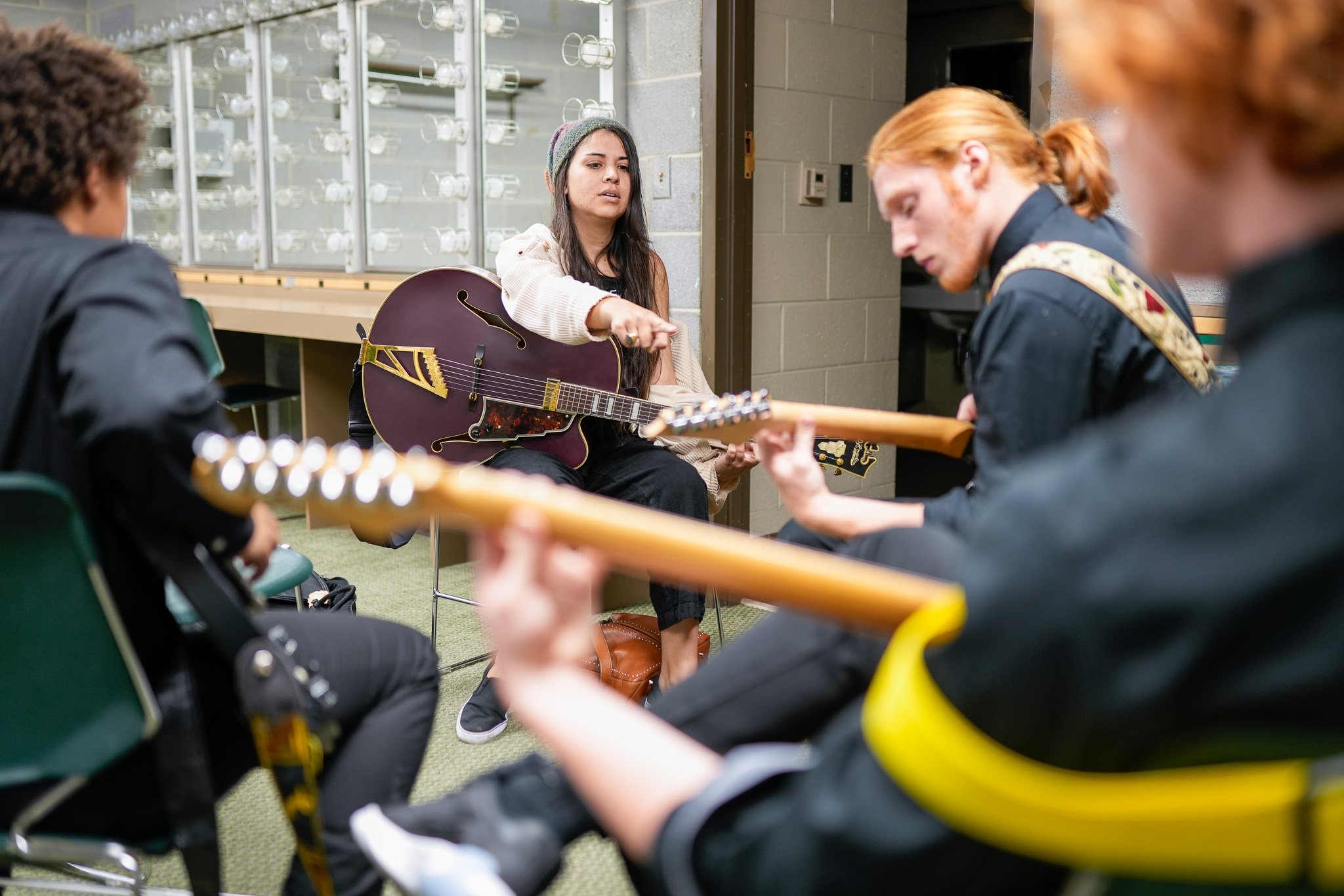 Three men holding guitars sit facing a woman who is seated at the front. The woman holds a purple guitar and gestures towards one of the guitars being held by the man seated on the right.