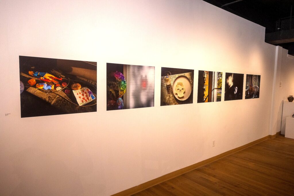 Six color photographs are mounted in a row on a white gallery wall. The images depict scenes of everyday life, including children's toys, a balloon, a plate with food shaped like a skull, and blurred portraits.