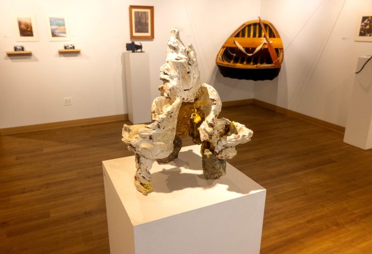 A ceramic sculpture with rough, textured white and gold surfaces is displayed on a pedestal in the gallery. Behind it, framed photographs and a suspended wooden coracle boat hang on the wall.