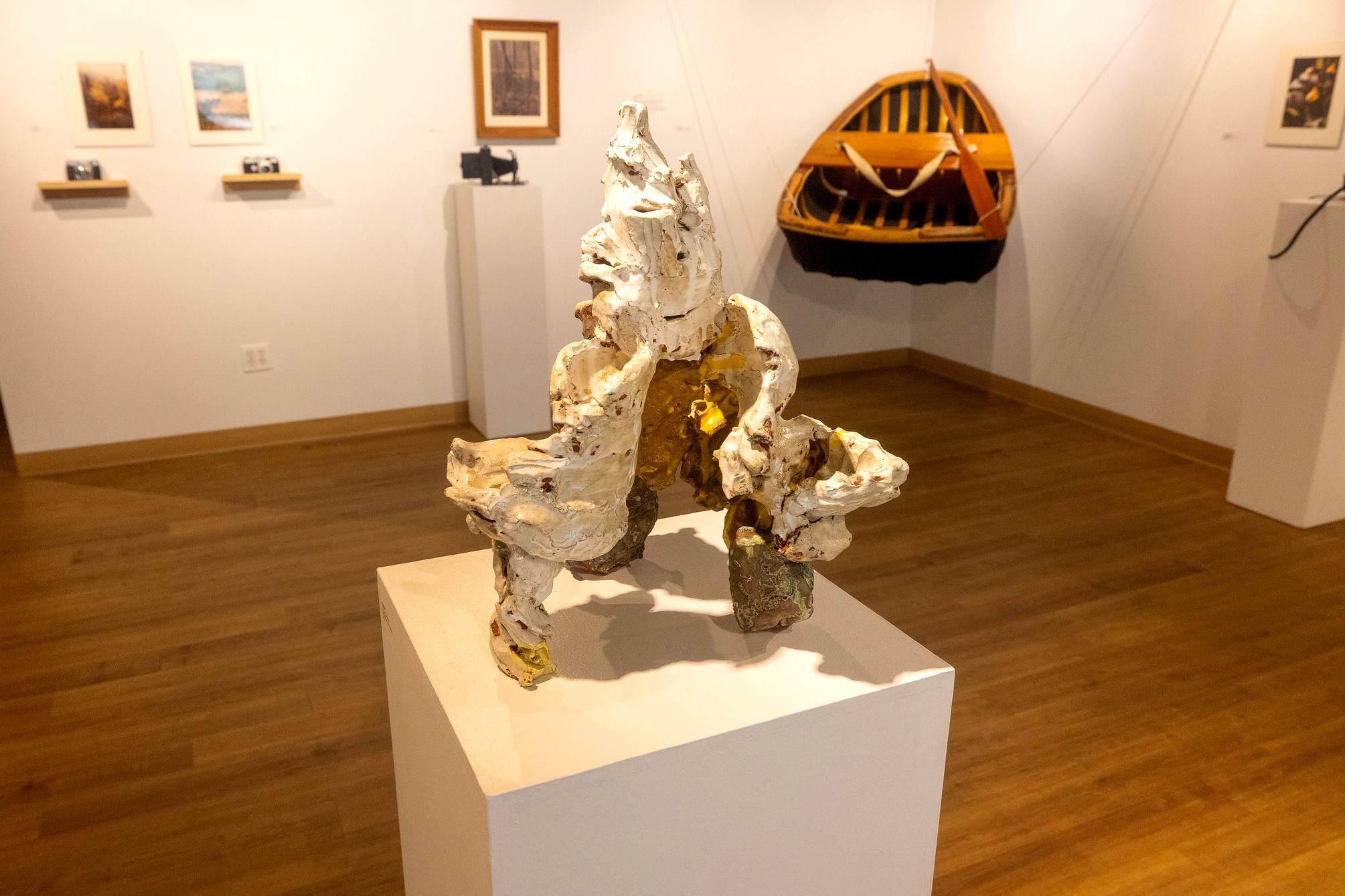 A ceramic sculpture with rough, textured white and gold surfaces is displayed on a pedestal in the gallery. Behind it, framed photographs and a suspended wooden coracle boat hang on the wall.