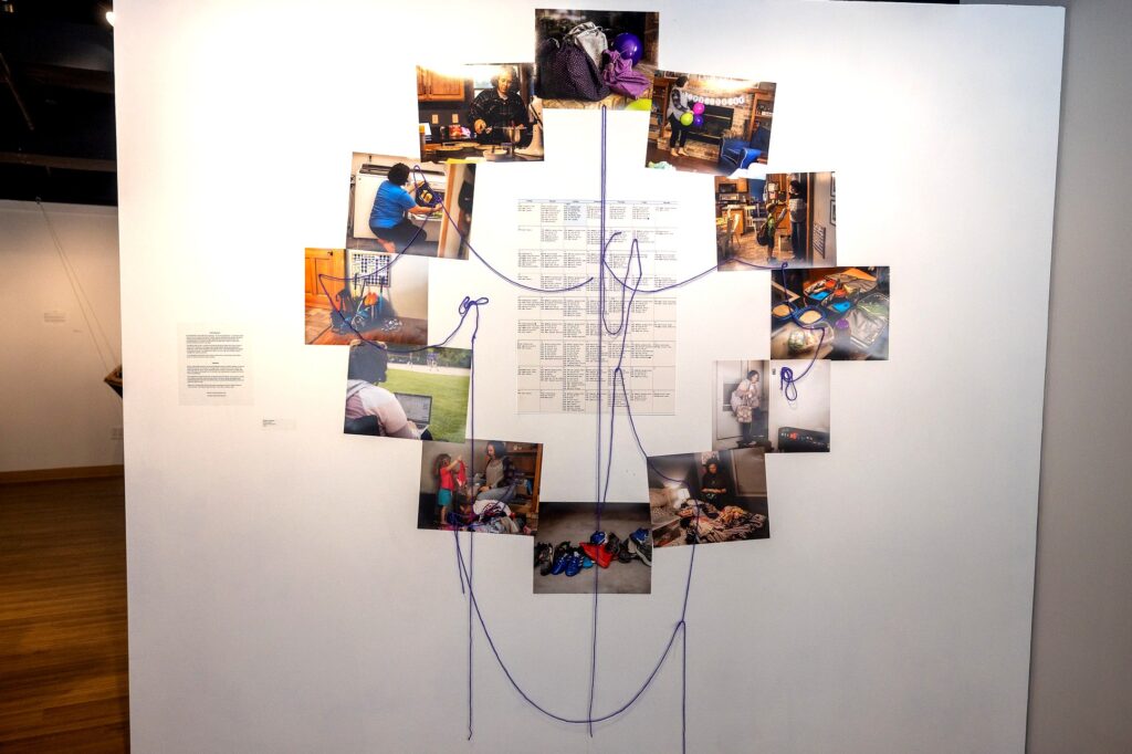 Wall installations of photographs connected by purple string surrounds a central calendar-like chart. The photos capture domestic and personal moments =, like cooking, parenting, working, and household tasks.