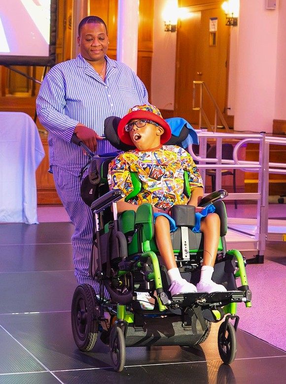 An adult pushes a child in a motorized wheelchair down a lit runway. The child wears a colorful patterned shirt, blue shorts, white shoes, and a red hat. The adult wears a blue-and-white striped outfit.