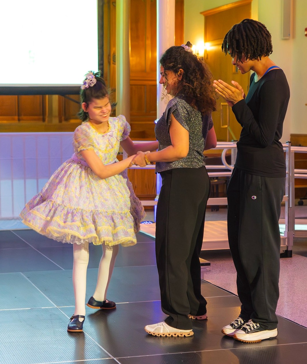 A young girl with a frilly lavender dress stands with another person in a glittery top and black pants, while a third person stands nearby clapping. They are on a runway.