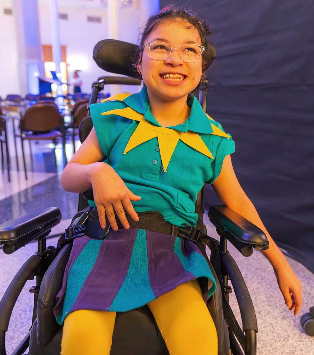 A smiling child in a wheelchair wears a bright teal and purple outfit with yellow tights and a star-shaped yellow collar. The background shows rows of chairs in a brightly lit event space.