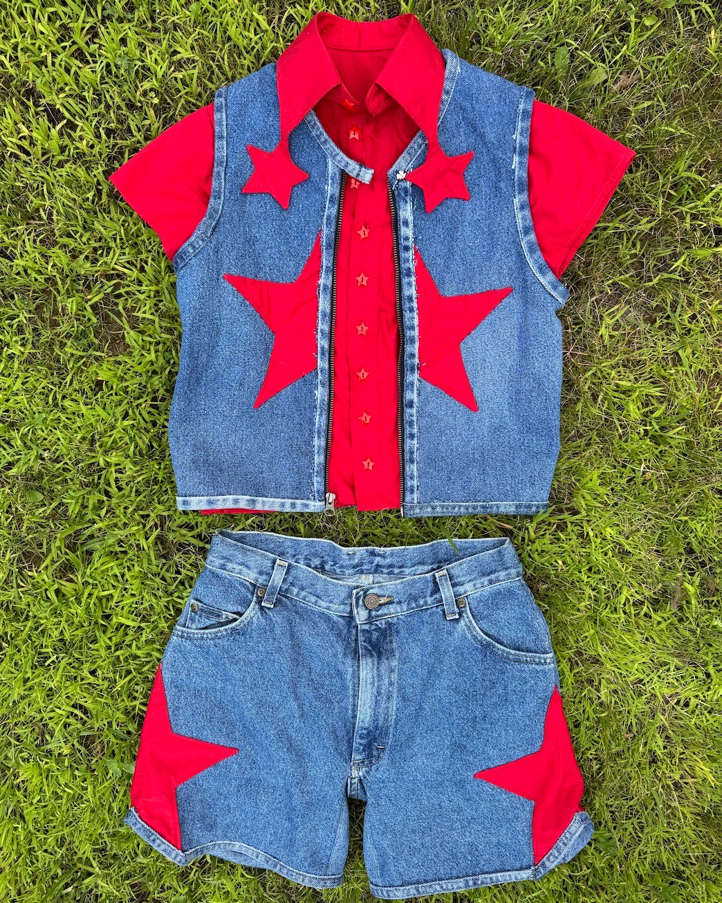 A denim vest and shorts with red star-shaped appliques and a red shirt underneath is laid flat on grass.
