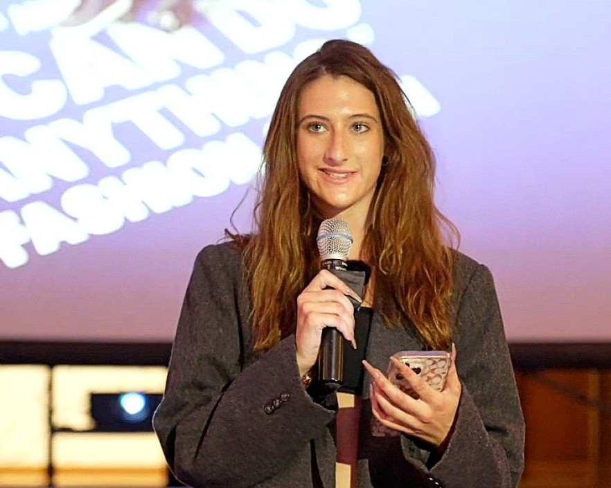A person holds a phone and microphone while speaking on stage, wearing a gray blazer and standing before a projection displaying the event's