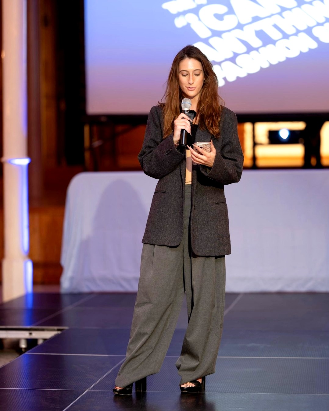 A person stands on a runway holding a microphone and phone, speaking to an audience. They wear a dark blazer, loose gray trousers, and open-toed heels. A large projection screen glows behind them with event text.