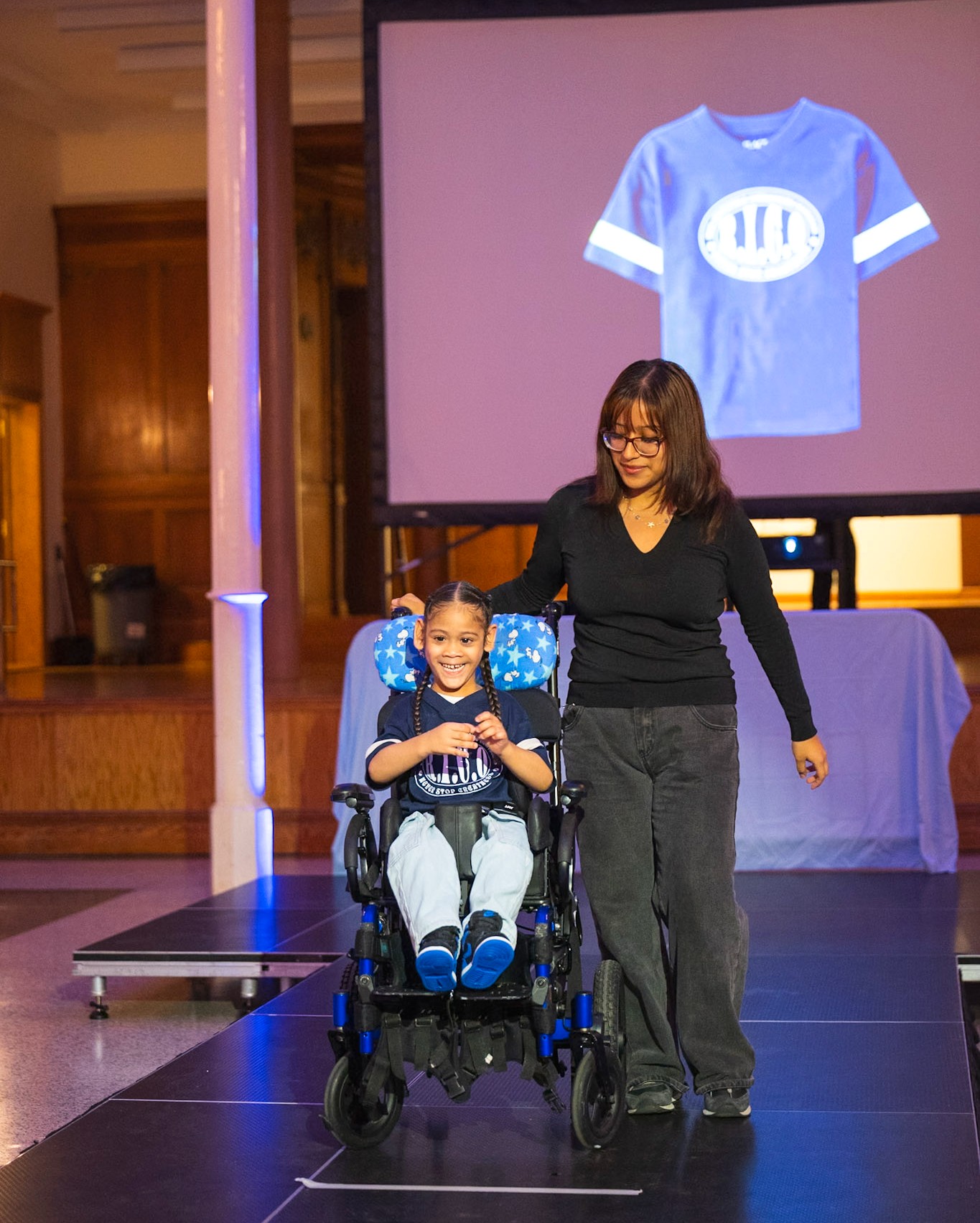 A child smiles while seated in a wheelchair on a runway, accompanied by an adult standing beside them. The child wears light blue jeans and a navy shirt with white accents. A projected image of the shirt is displayed behind them.