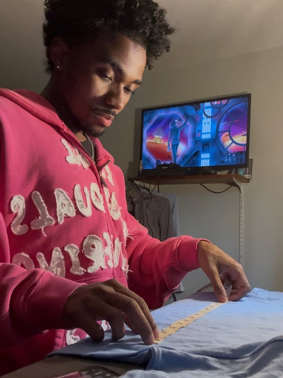 A person wearing a bright pink hoodie with white text concentrates while measuring fabric on a table. A television in the background shows an animated scene.