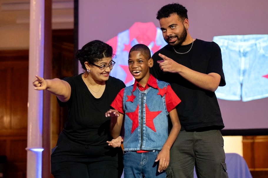 A smiling child wearing a denim vest with a large red star stands between two adults dressed in black. All three point and smile toward the audience on a brightly lit runway.