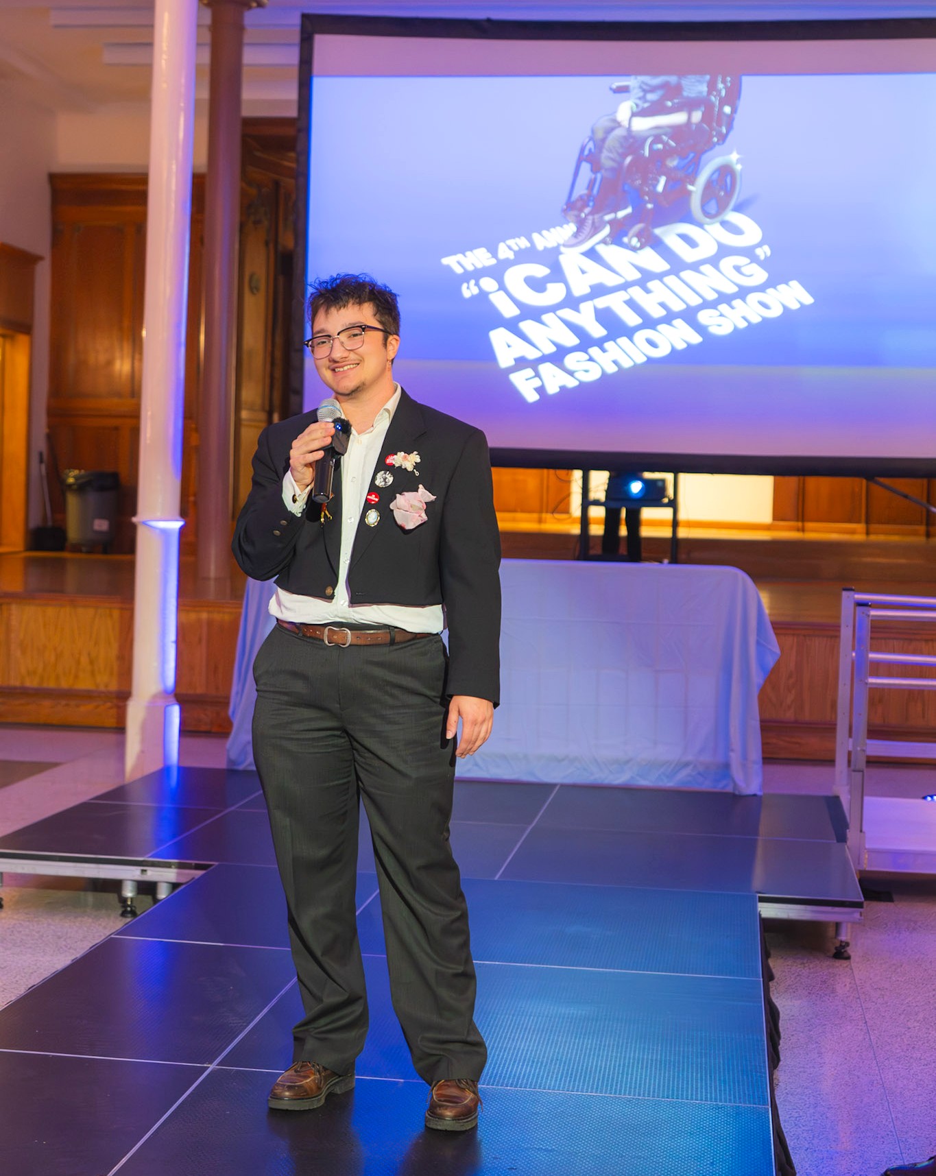 A person stands holding a microphone and smiling. They wear a dark suit with a white shirt and decorative pins and flowers on the jacket. A large projection behind the reads