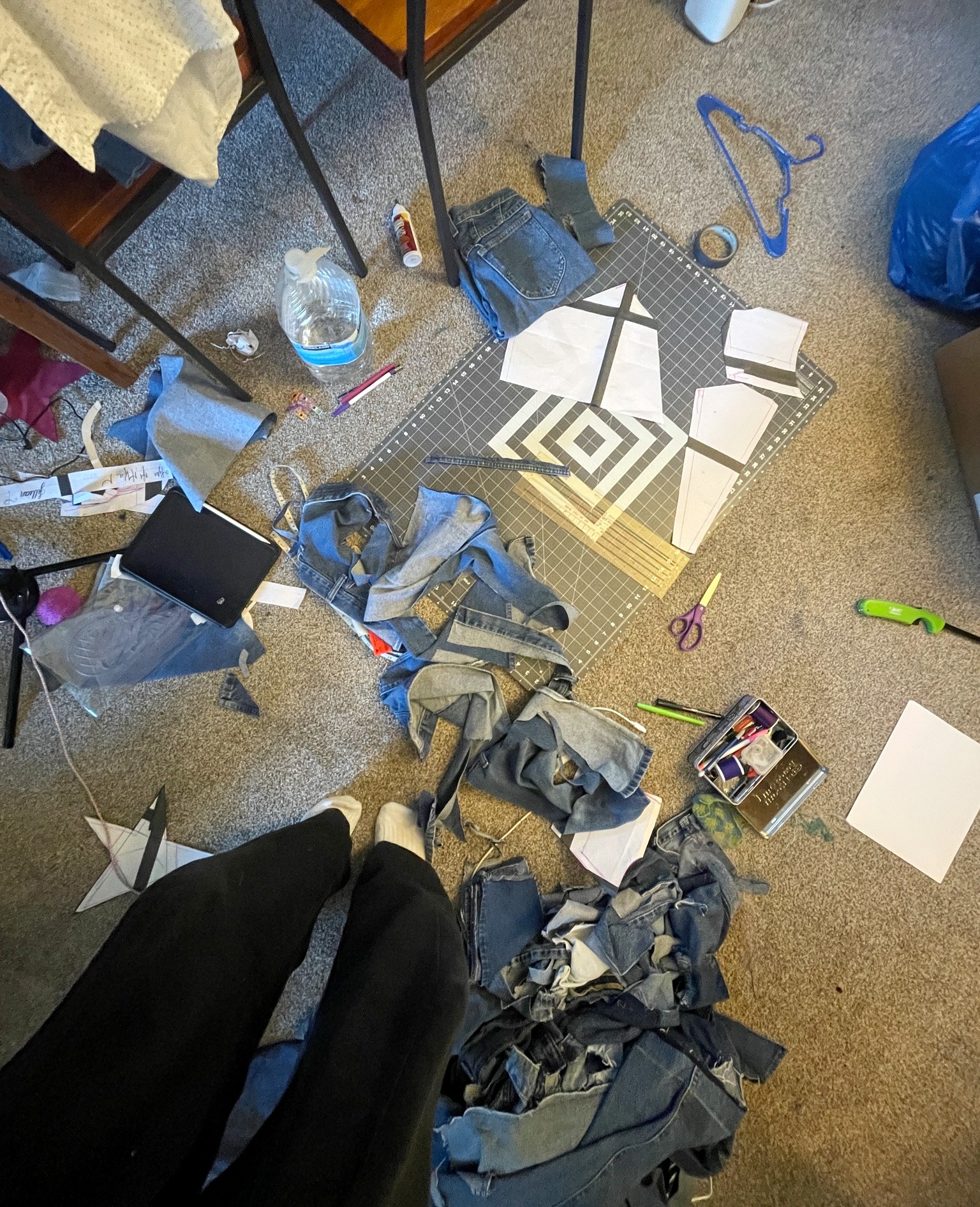 A workspace floor covered in denim fabric scraps, paper patterns, scissors, a cutting mat, and sewing supplies. A person's feet in socks and black pants are visible, standing among the materials.