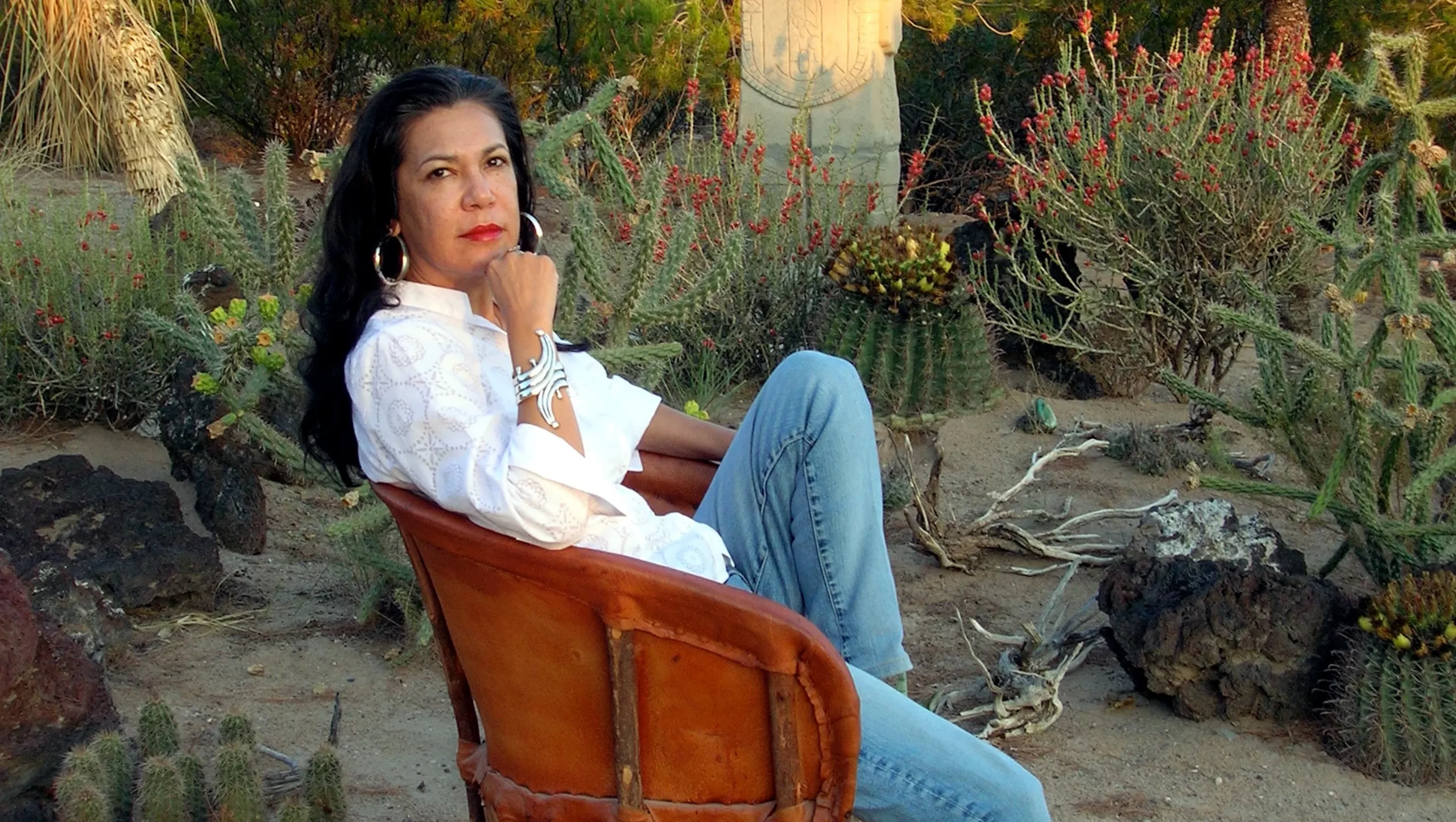Woman sits in white shirt and jeans on suede chair surrounded by cacti