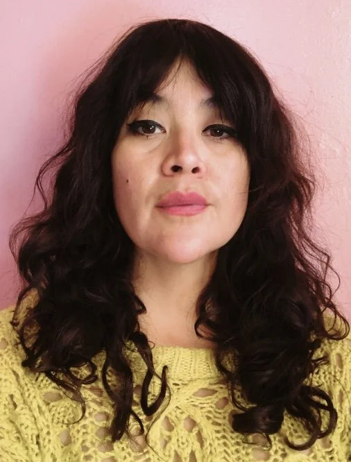 Woman with medium length wavy black hair and yellow crochet shirt looks at camera in front of pink wall