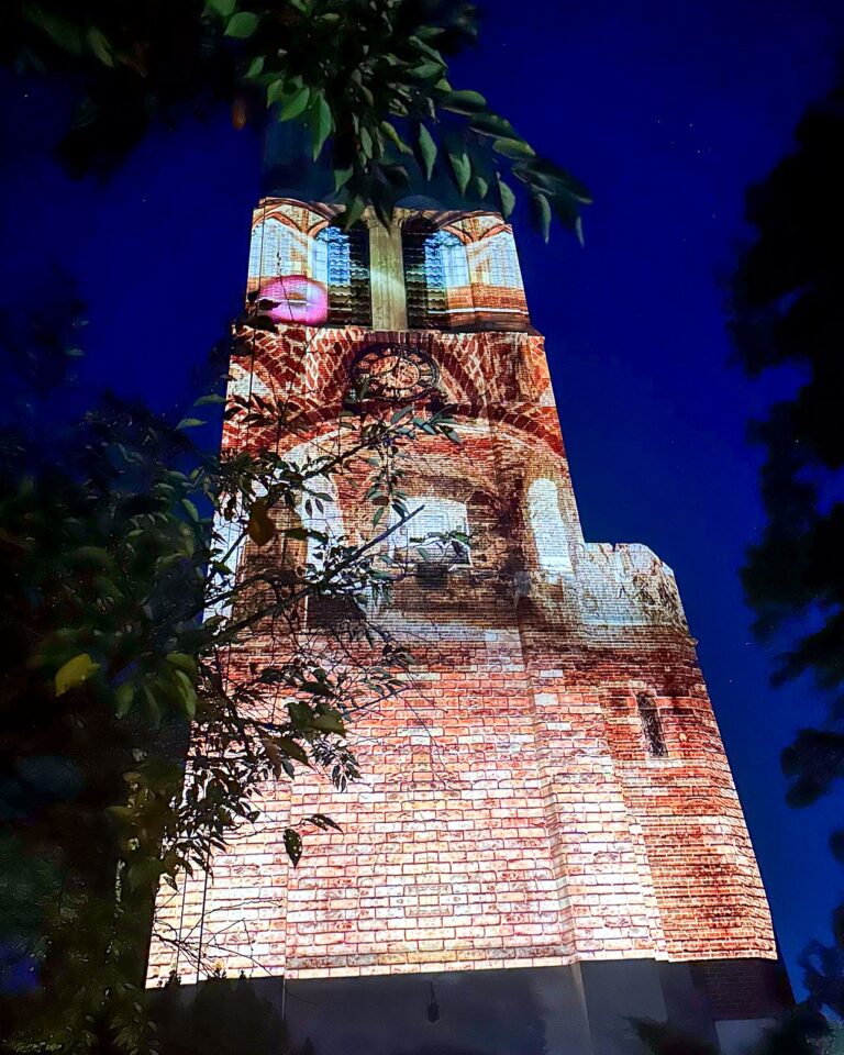 A nighttime view of a tall brick clock tower partially framed by leafy branches, illuminated with a warm-toned light projection that highlights its arched windows and brickwork against a clear night sky.