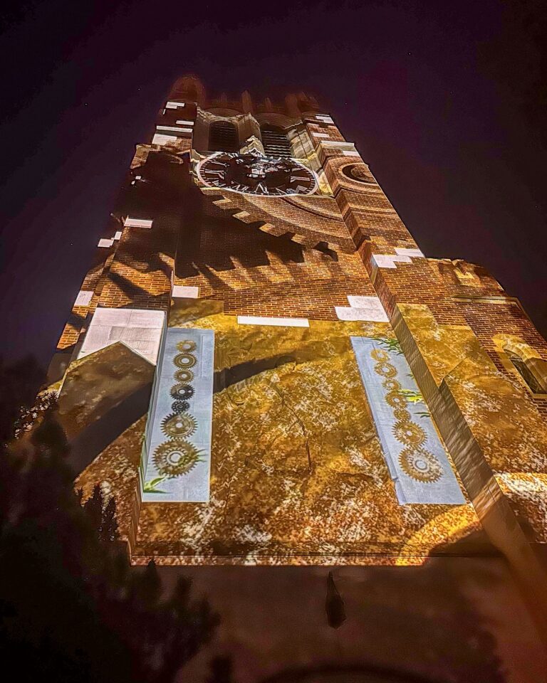 Beaumont Tower illuminated in warm golden tones with projected imagery of turning gears and clockwork patterns extending upwards toward the clock face.