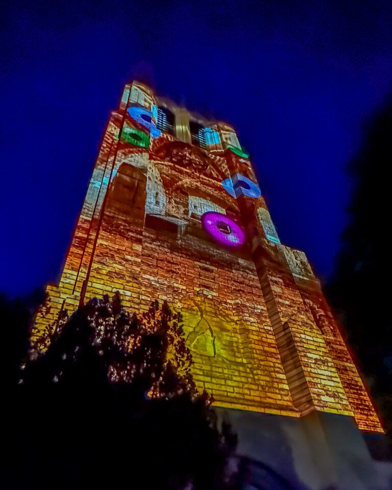 A clocktower glows with vibrant, multicolored light projections featuring geometric shapes and circular patterns in blue, pink, green, and orange.